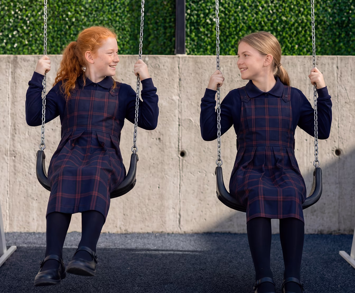 Two young girls in plaid school uniforms sitting on swings, smiling and looking at each other outdoors.