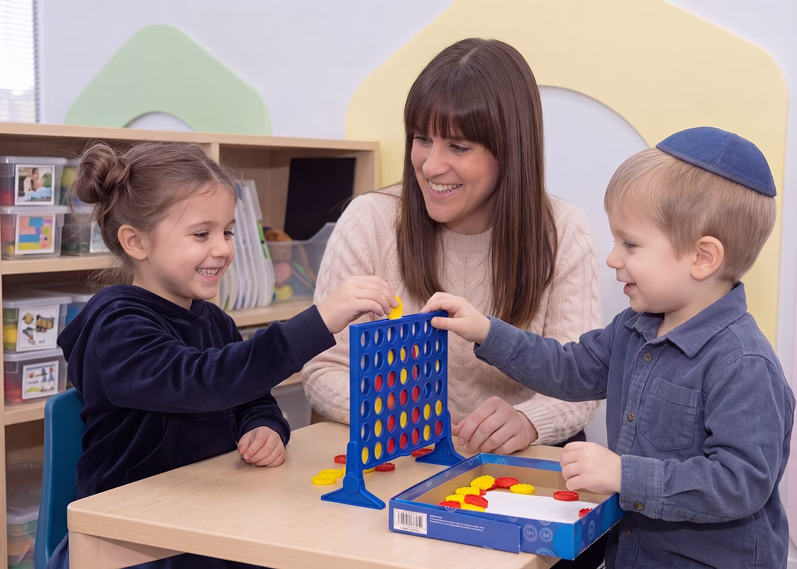 Two young children playing Connect Four at a table with a school therapist watching them.
