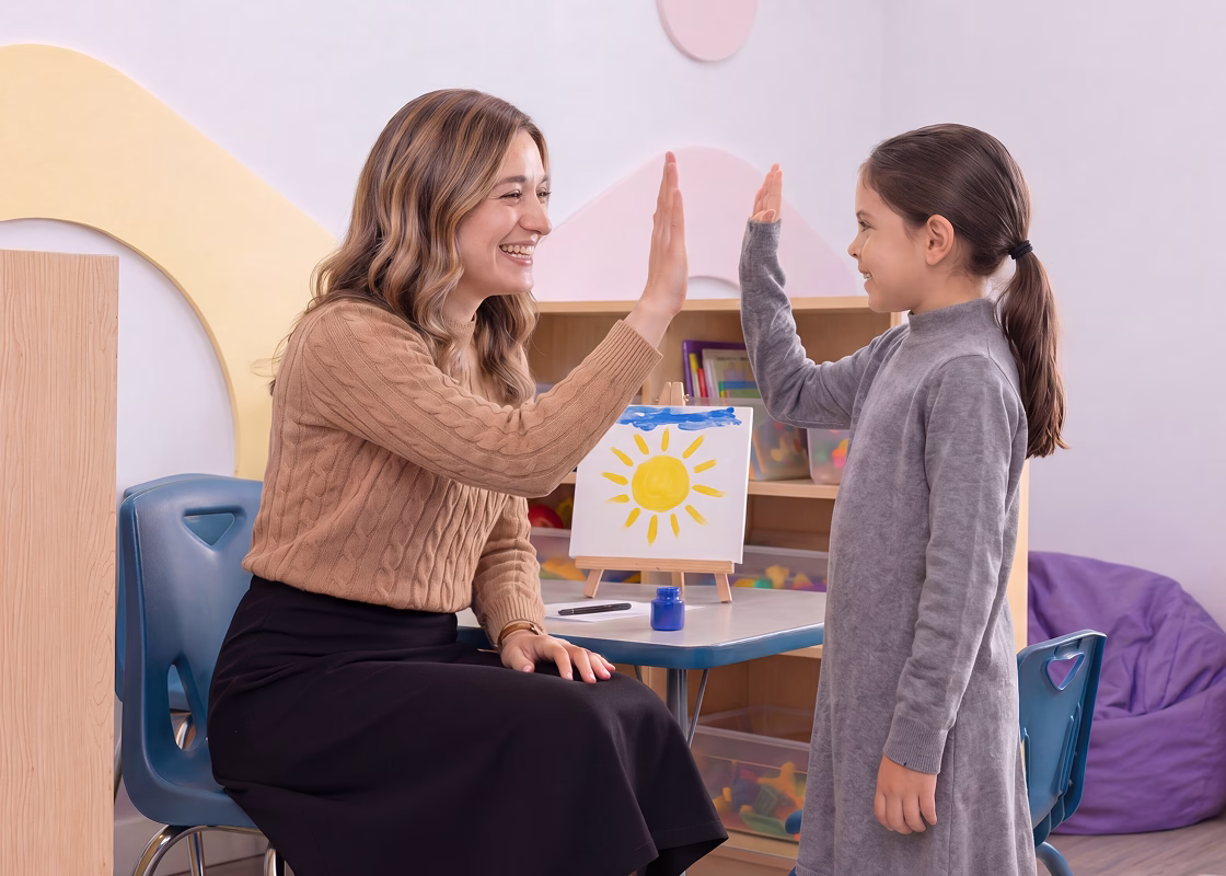 Smiling therapist and little girl giving each other a high five in a classroom with a sun painting on the table.