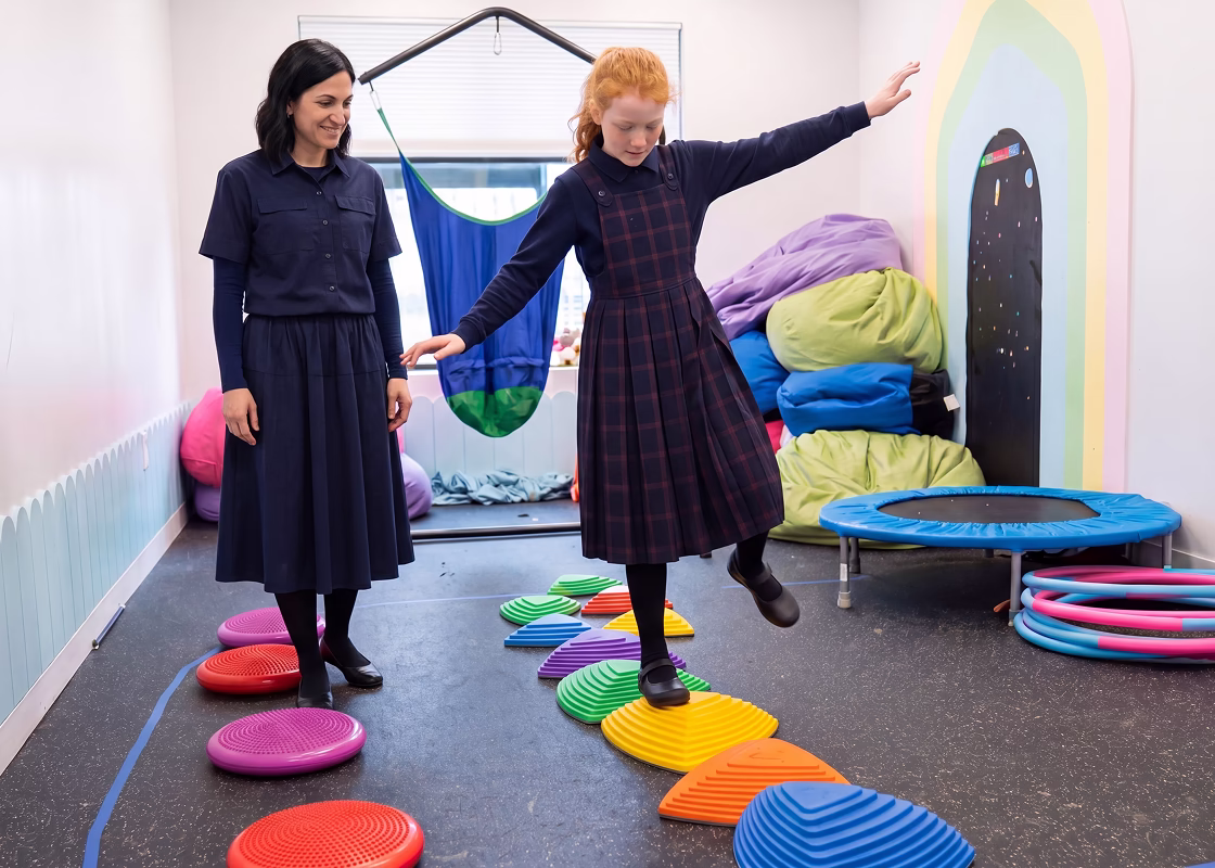 Girl balancing on colorful stepping stones while a therapist watches in a room with more equipment.