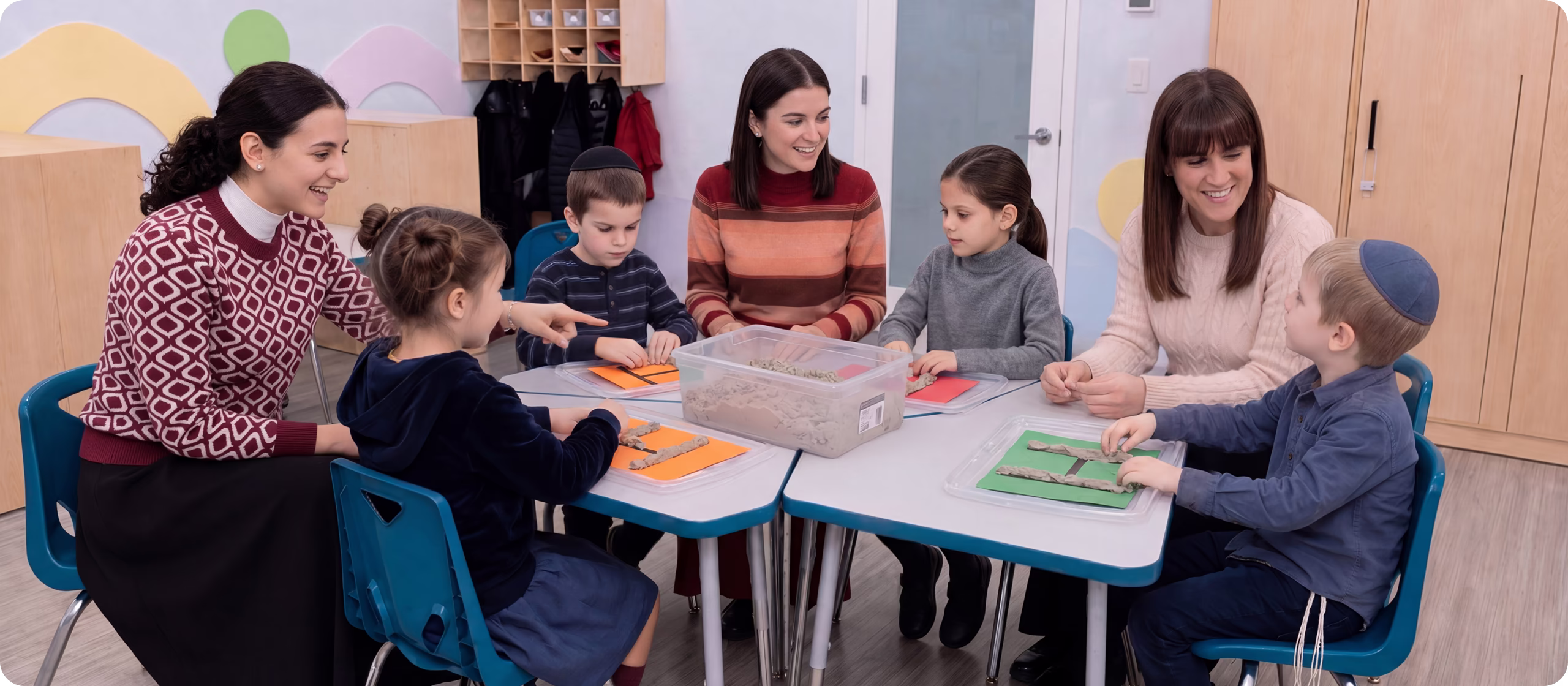 Three teachers and four children sitting around a table in a classroom working with sand to create the letter H on a colored paper.