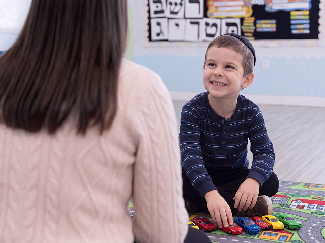 Smiling boy sitting on play mat with toy cars, facing a teacher in a classroom setting.