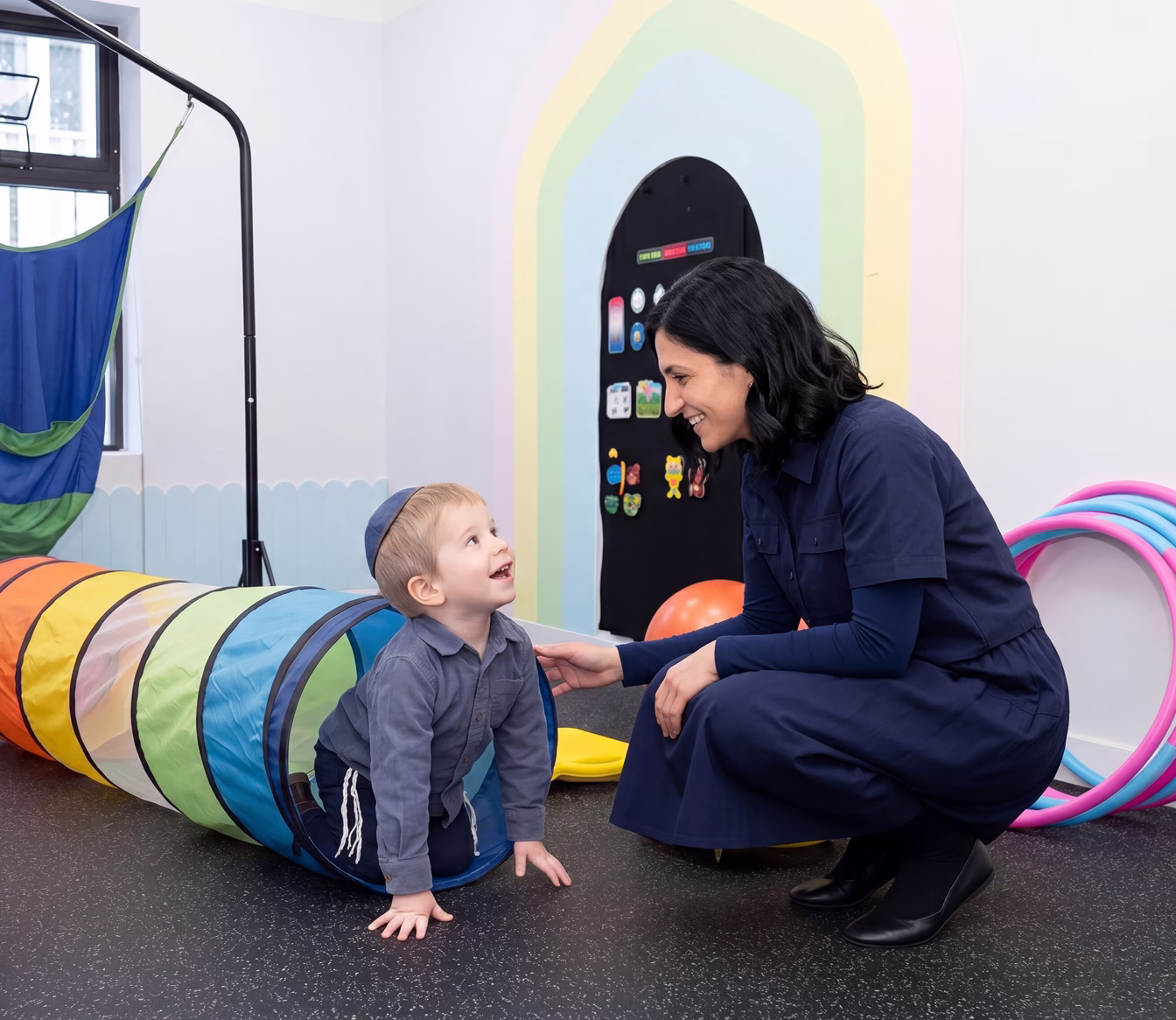 Smiling therapist crouches near a boy crawling out of a colorful play tunnel in a brightly lit room.