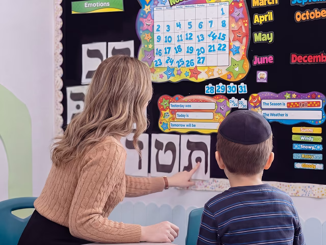 Teacher pointing to a Hebrew letter on a bulletin board with a little boy watching.