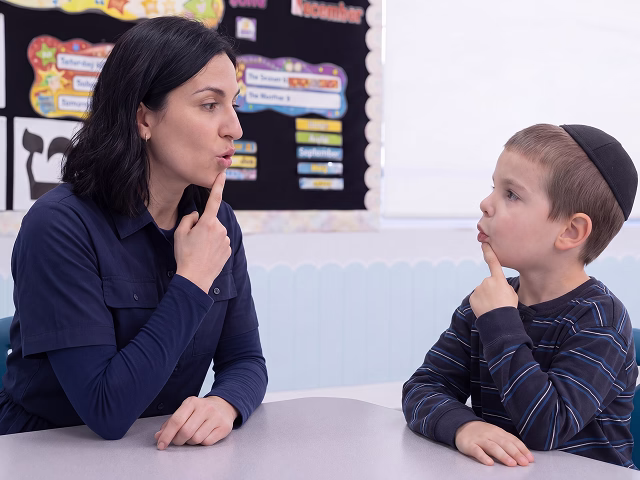 Teacher and young boy using finger to lips gesture in classroom.