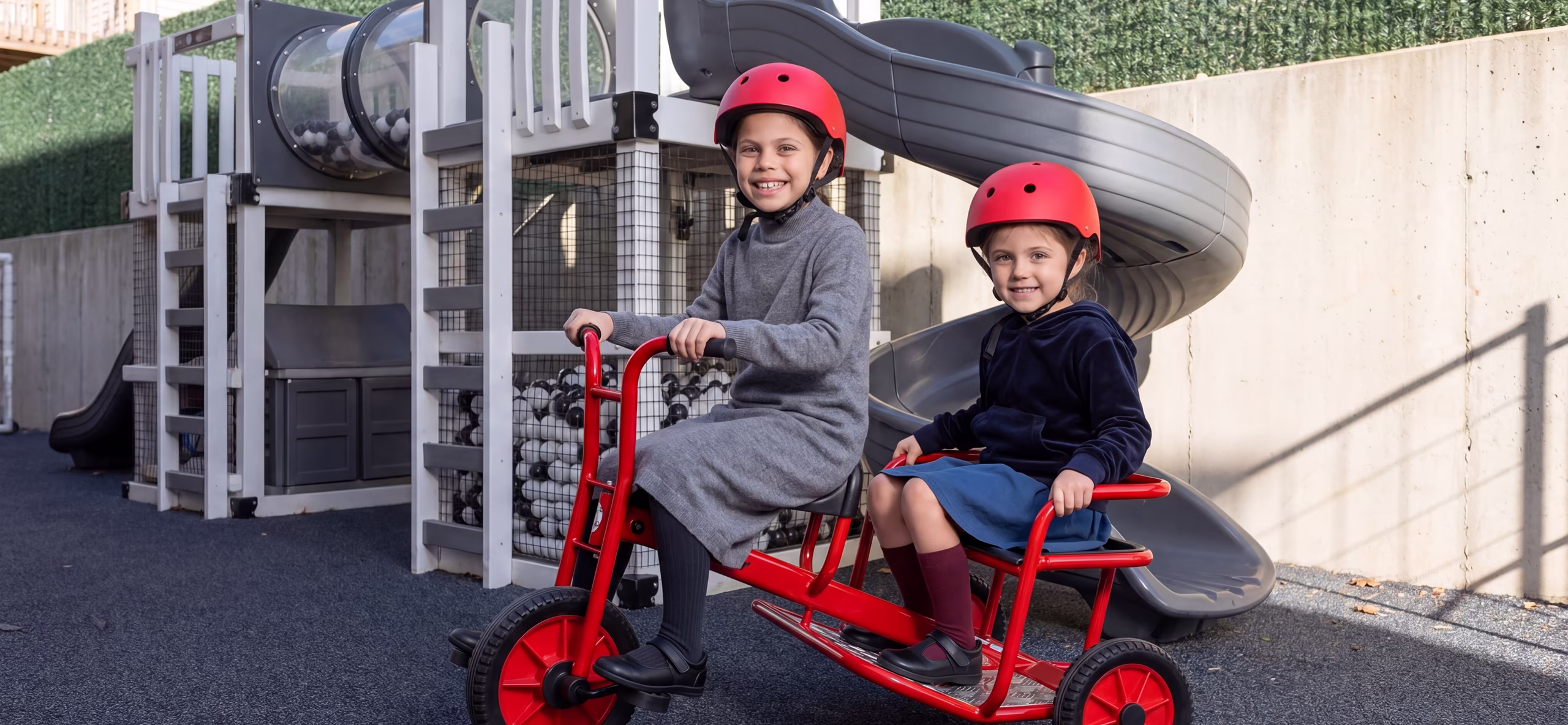 2 young girls on a red bike outdoors by a playground.