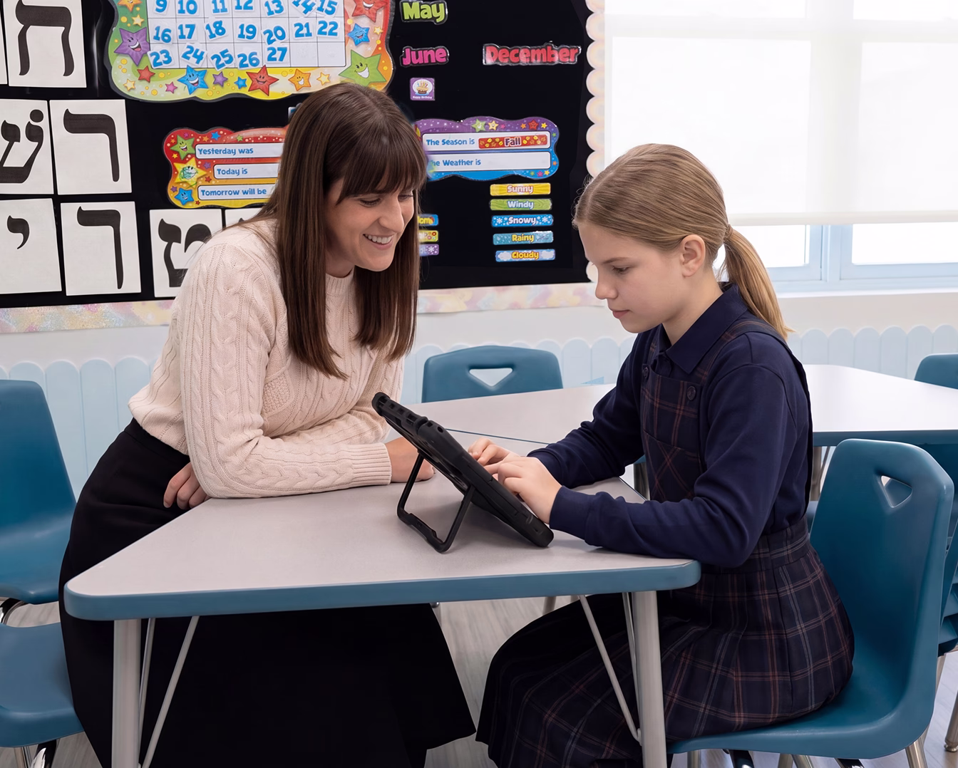A young girl using her AAC device while sitting at a table in a classroom with her teacher watching her.