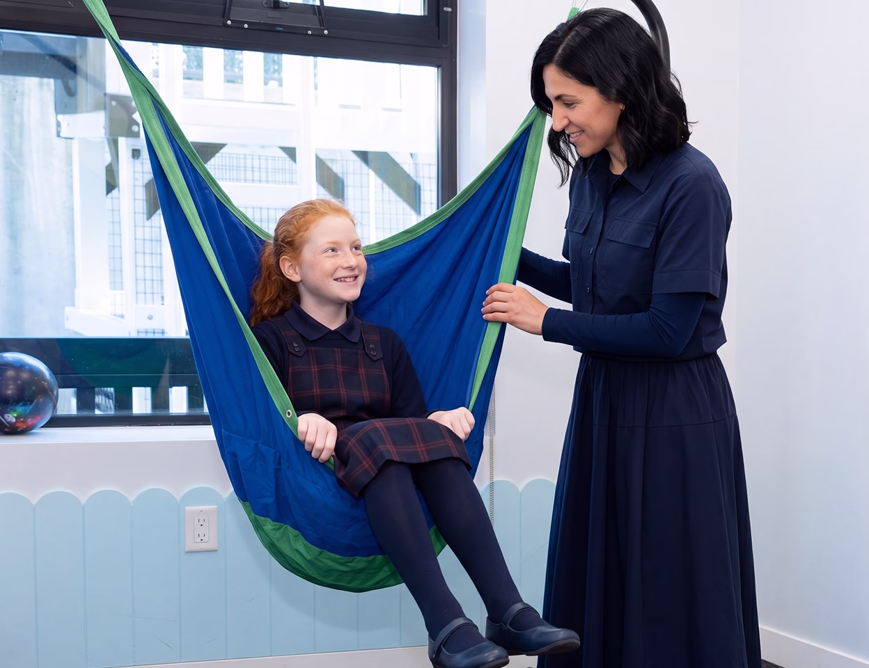 A young girl in a therapeutic hammoc with her teacher looking at her smiling.