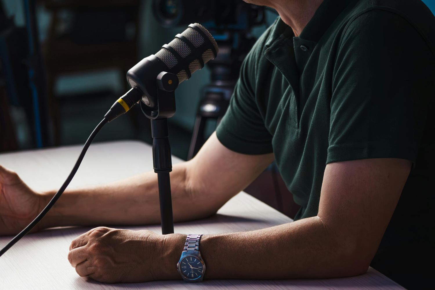 Person wearing a watch speaking into a microphone on a table, with a camera blurred in the background.
