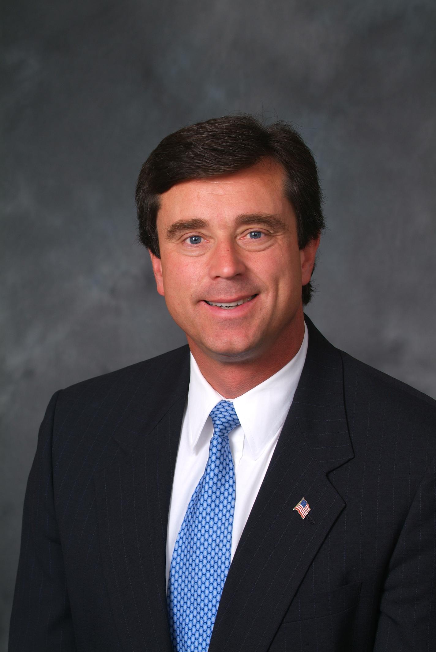 Smiling man with dark hair wearing a dark suit, white shirt, blue tie, and US flag pin.