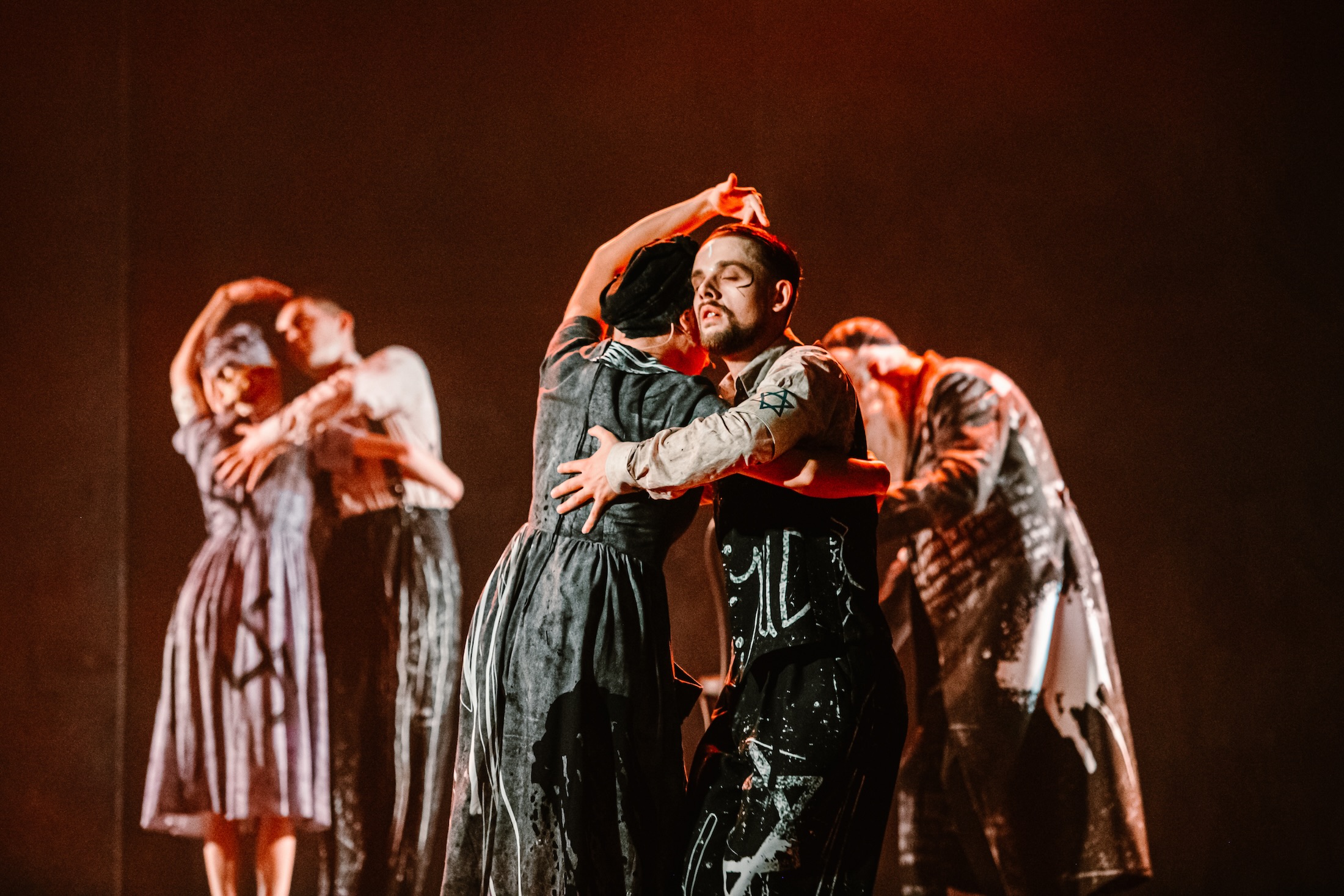 Two performers embrace as figures behind them mirror the gesture in a warmly lit scene from Irena.