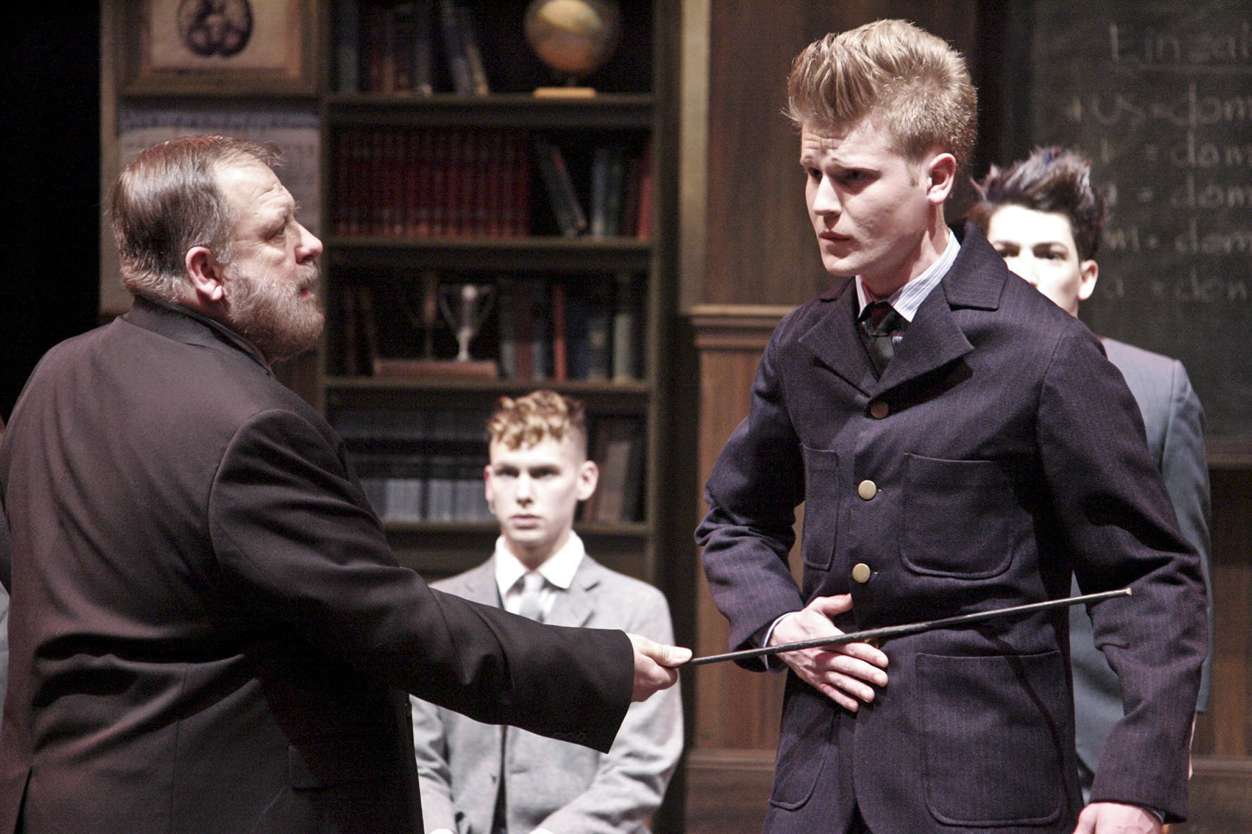An adult man confronts a young man while other students watch in a classroom scene from Spring Awakening.