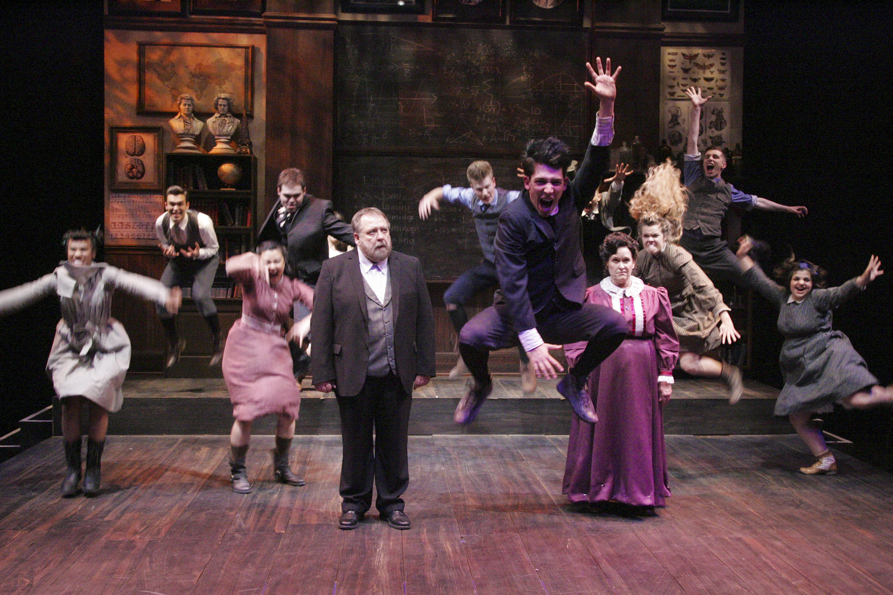 The Spring Awakening ensemble erupts in movement around two rigid adults in a classroom.