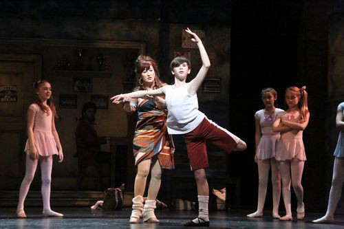 Mrs. Wilkinson guides Billy through a ballet position as young dancers watch in Billy Elliot.