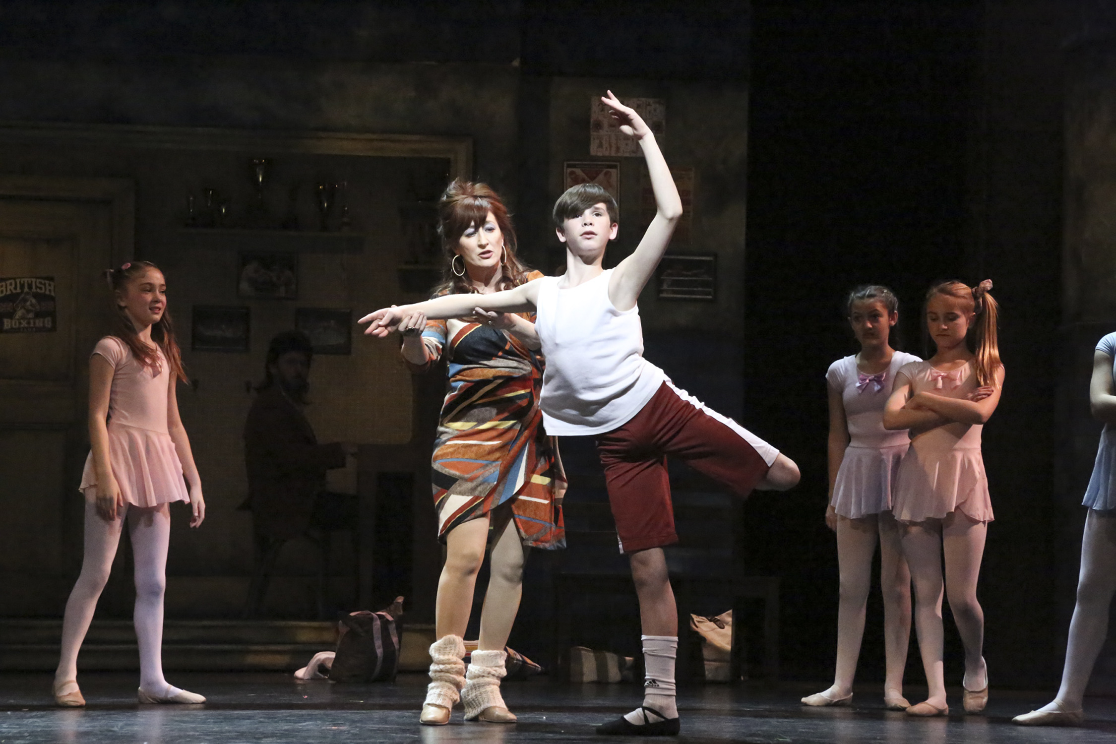 Mrs. Wilkinson guides Billy through a ballet position as young dancers watch in Billy Elliot.