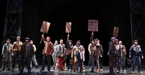 An ensemble of striking miners and families holds protest signs across the stage in Billy Elliot.
