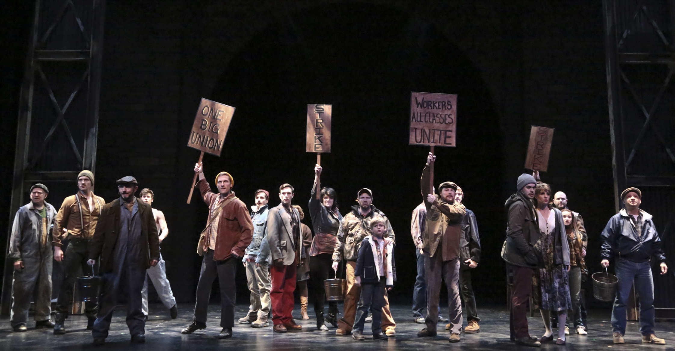 An ensemble of striking miners and families holds protest signs across the stage in Billy Elliot.