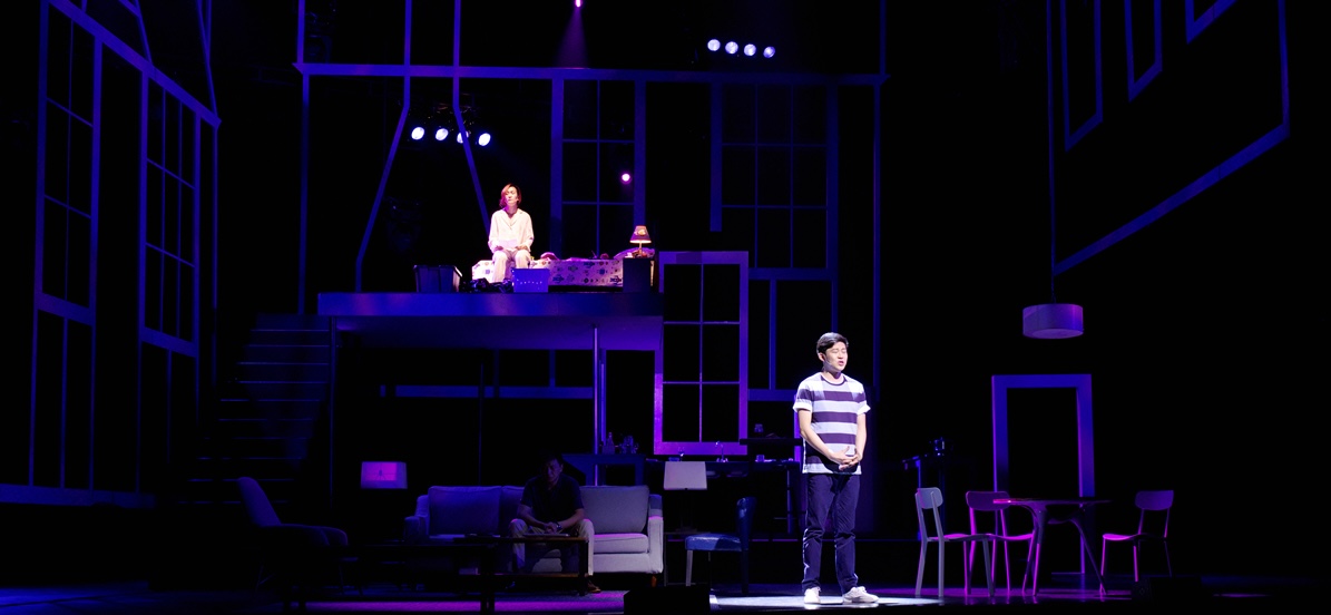 Jason stands in a pool of light below Becca on an elevated bedroom platform, while Howie sits in shadow on the couch in Rabbit Hole.