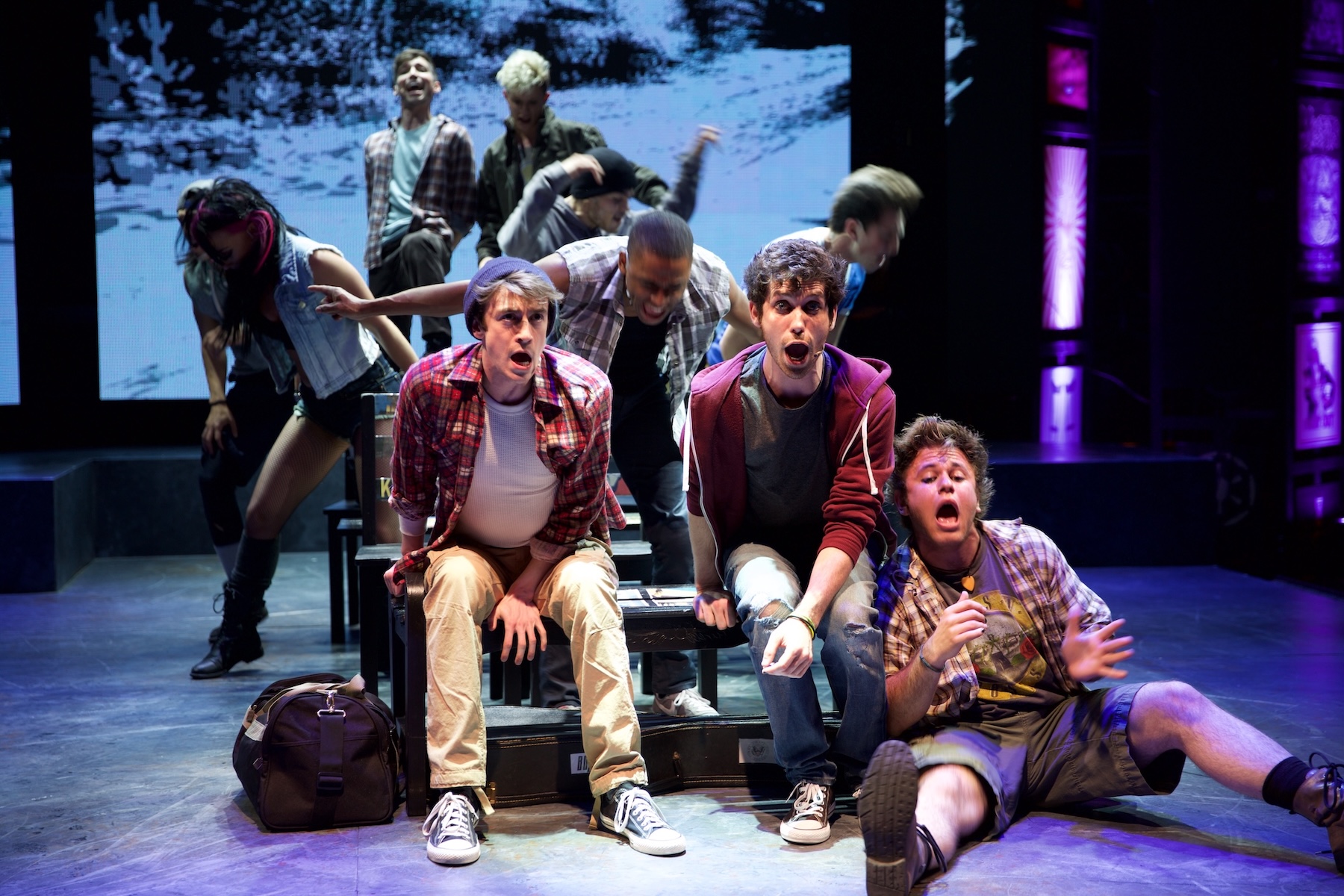 Three young men ride an imaginary bus as the ensemble gathers behind them in American Idiot.