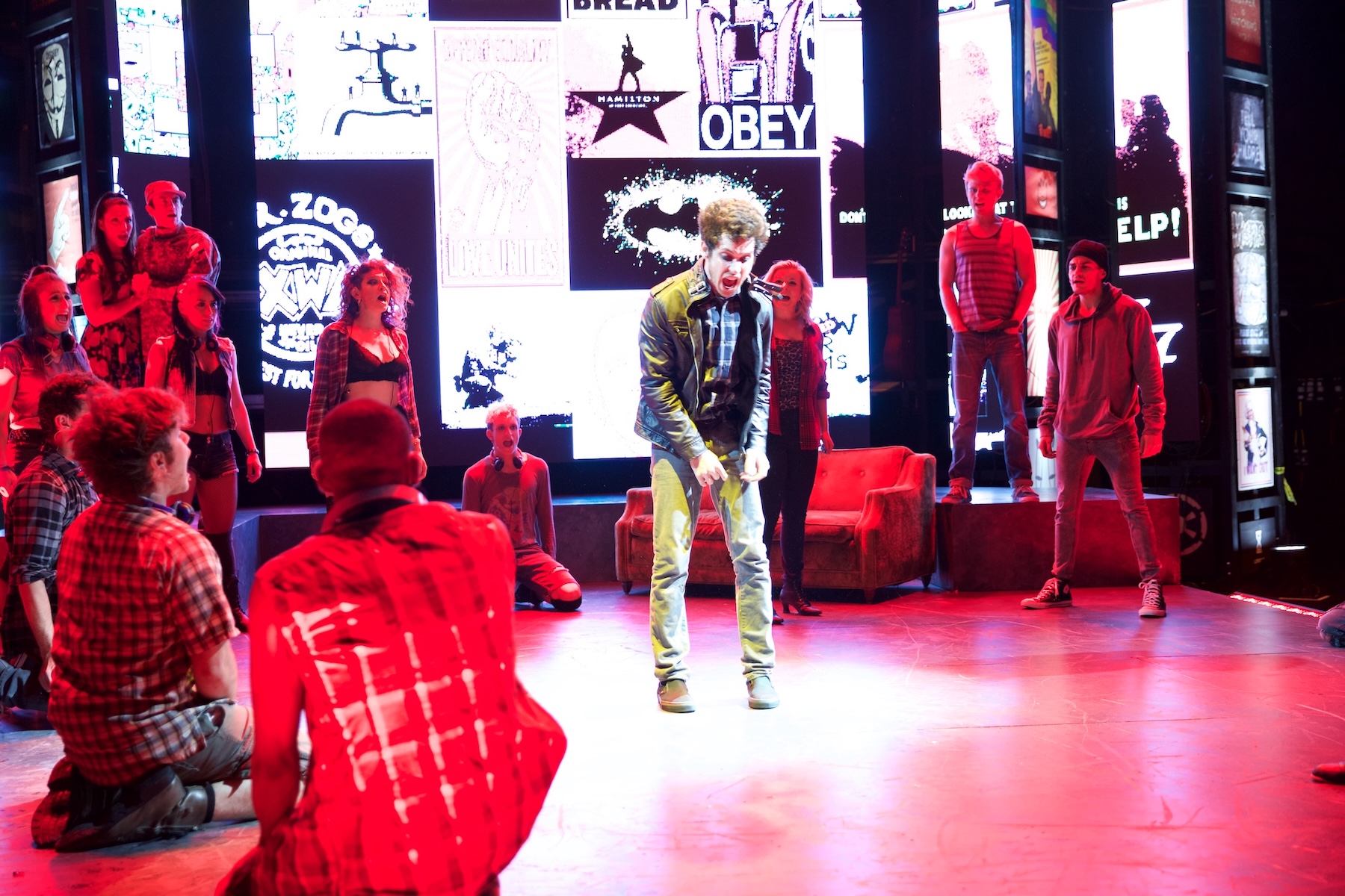 A young man stands alone amid a red-lit ensemble and projected media imagery in American Idiot.
