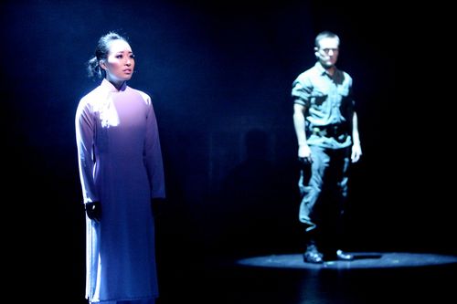 Kim stands in a white áo dài as Chris appears behind her in blue stage light in Miss Saigon.