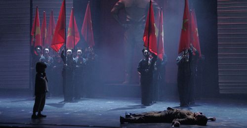 Tam stands before masked soldiers holding red flags as Thuy lies on the ground in Miss Saigon.