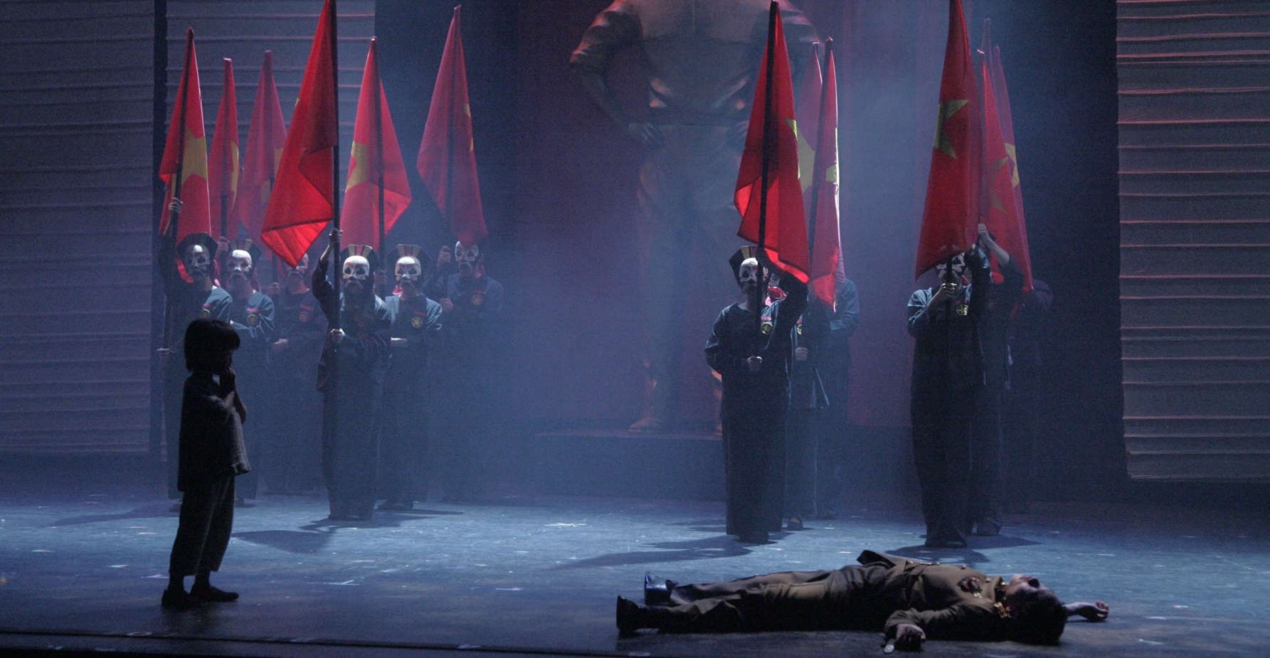 Tam stands before masked soldiers holding red flags as Thuy lies on the ground in Miss Saigon.
