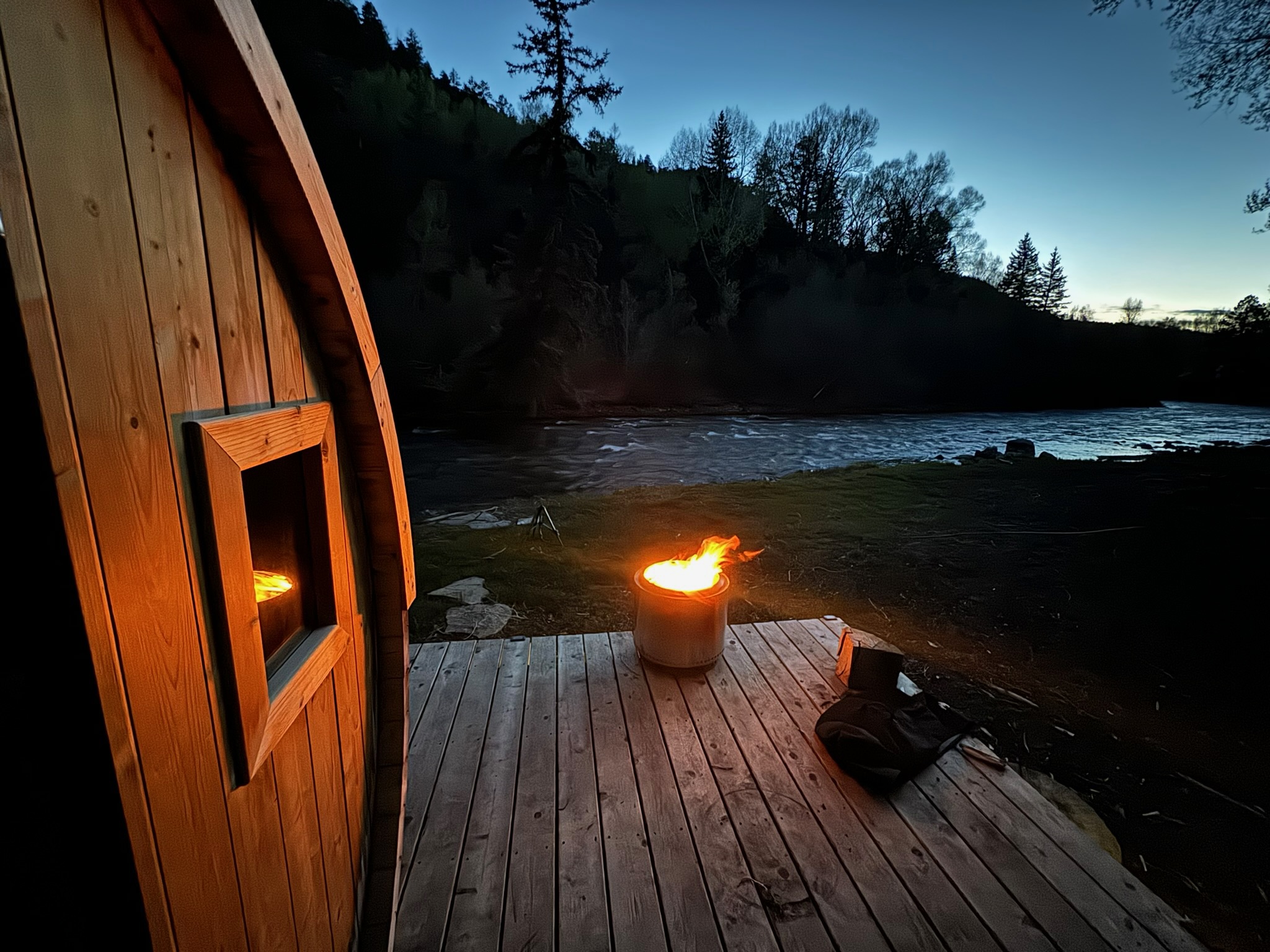 Barrel sauna by the river at dusk with fire pit