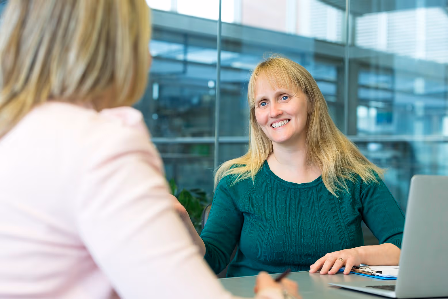 A woman with blonde hair wearing a green shirt, sitting at desk with a colleague whose back is facing the camera