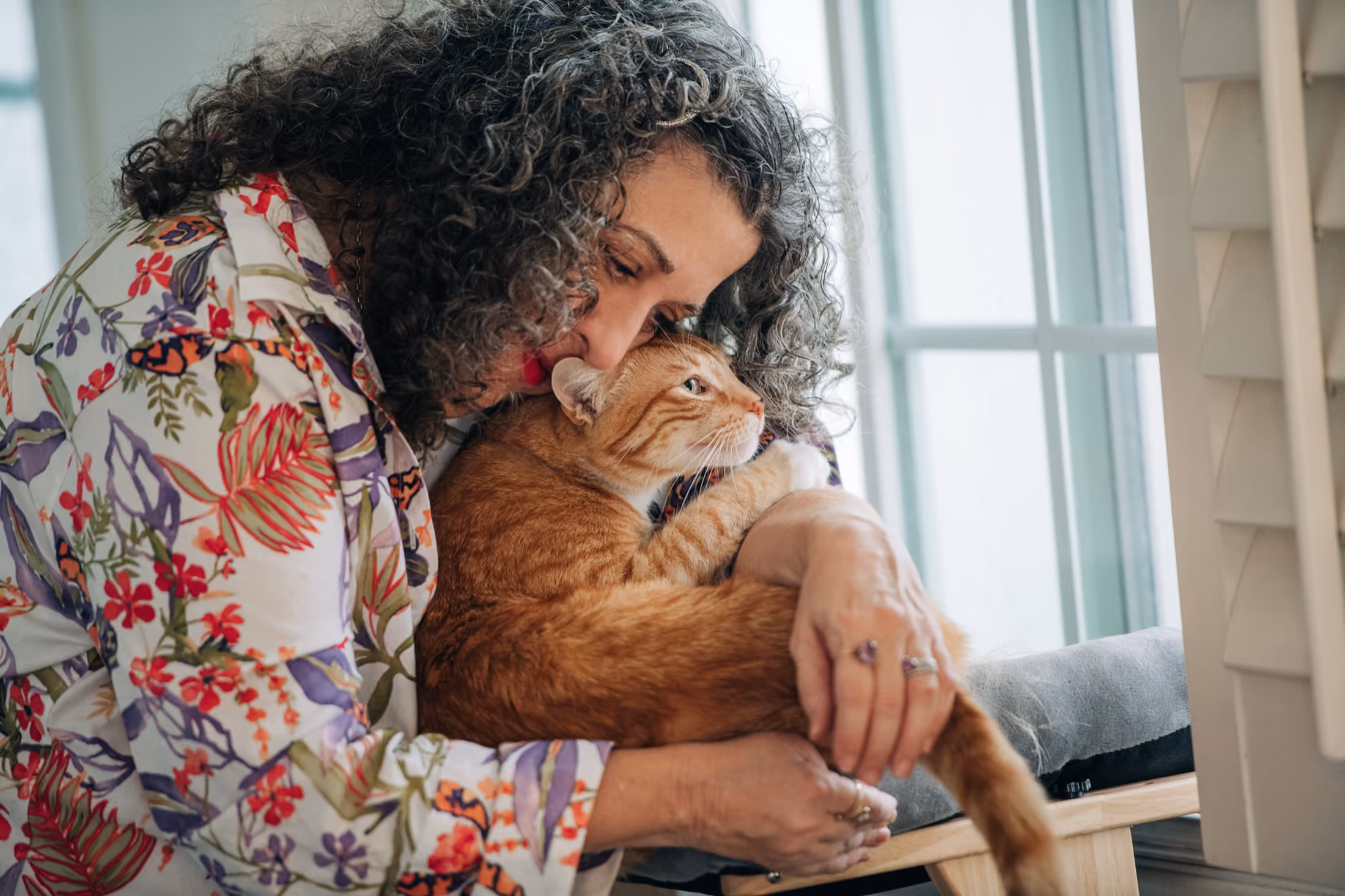 Kara hugging and kissing an orange tabby cat near a window.