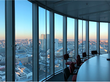 View of a city skyline at sunset through tall curved glass windows in a modern office conference room with chairs around a table.