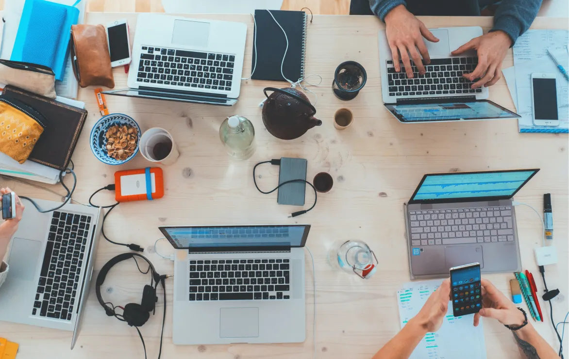 Overhead view of a wooden table with four laptops, smartphones, notebooks, headphones, a teapot, snacks, and hands typing and holding a phone.