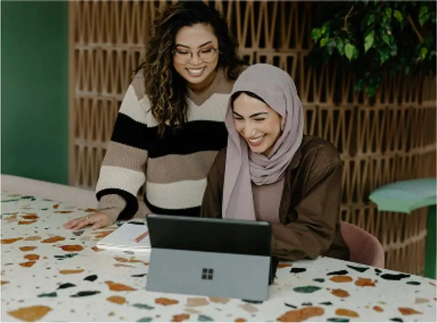 Two women smiling and looking at a laptop computer on a terrazzo table in a cozy modern setting.