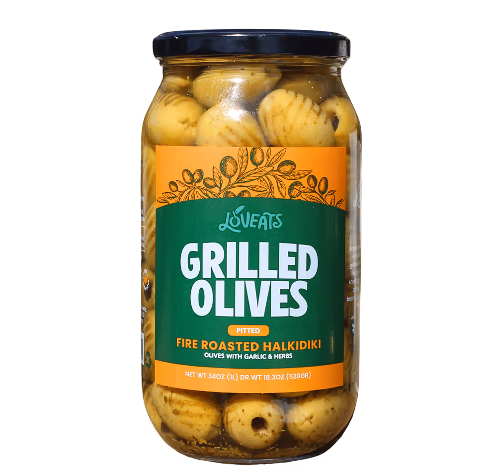 Front of Jar of Loveats grilled olives. Garlic head cut it half as well as some garlic cloves, fresh oregano and olive tree small branches in the background.