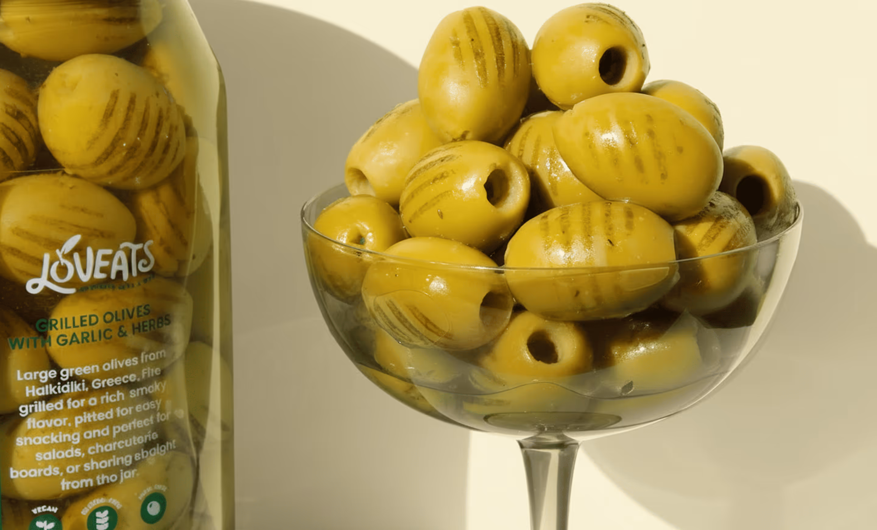 Grilled green olives piled up in a coupe glass next to olive jar.