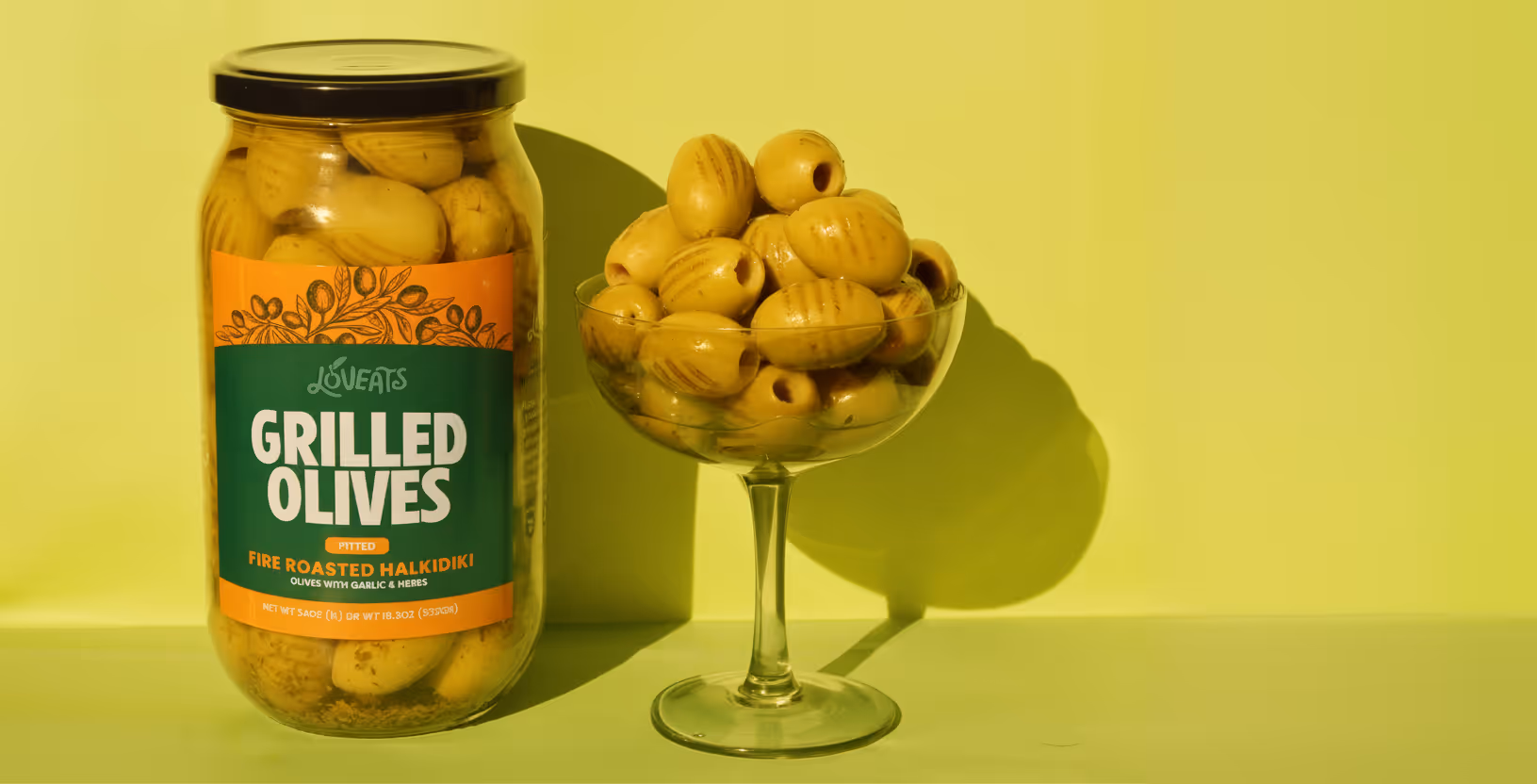 Jar of fire roasted Halkidiki grilled green olives next to a glass filled with the same olives.