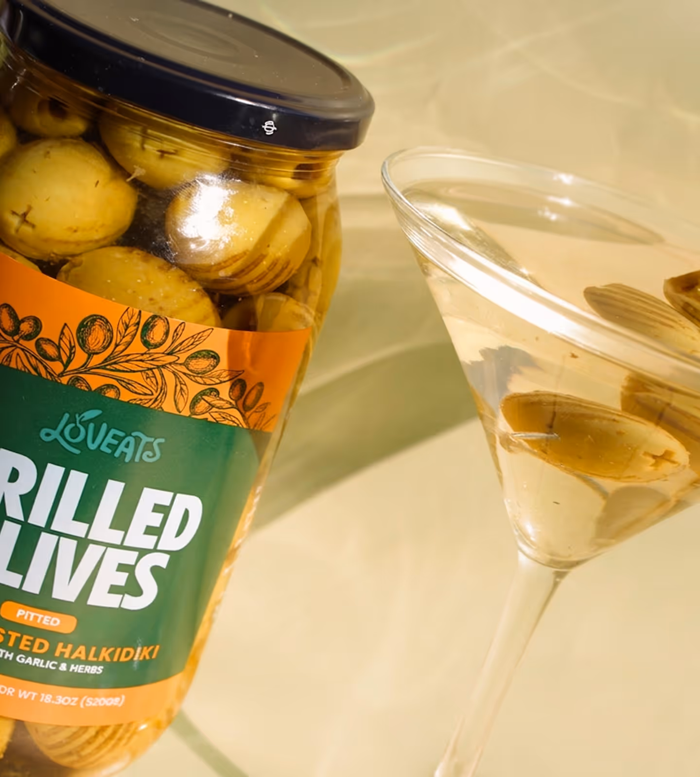 Jar of pitted grilled Halkidiki olives next to a clear cocktail glass with olives inside.
