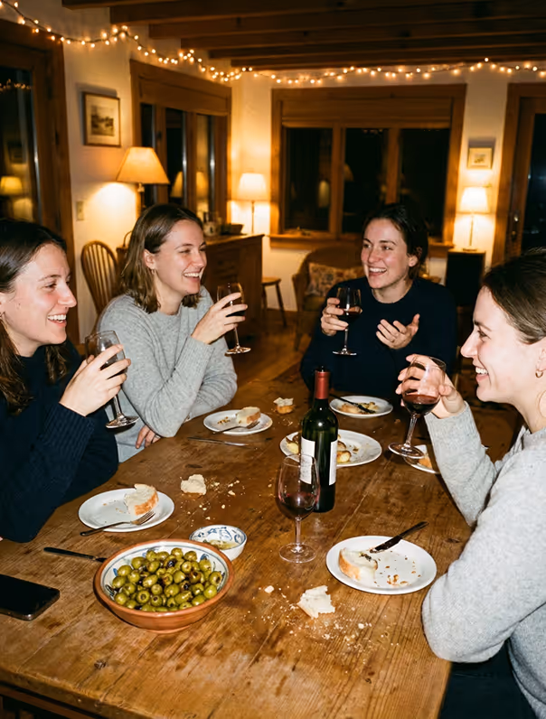 Four women laughing and drinking wine around a wooden table with olives and bread crumbs.