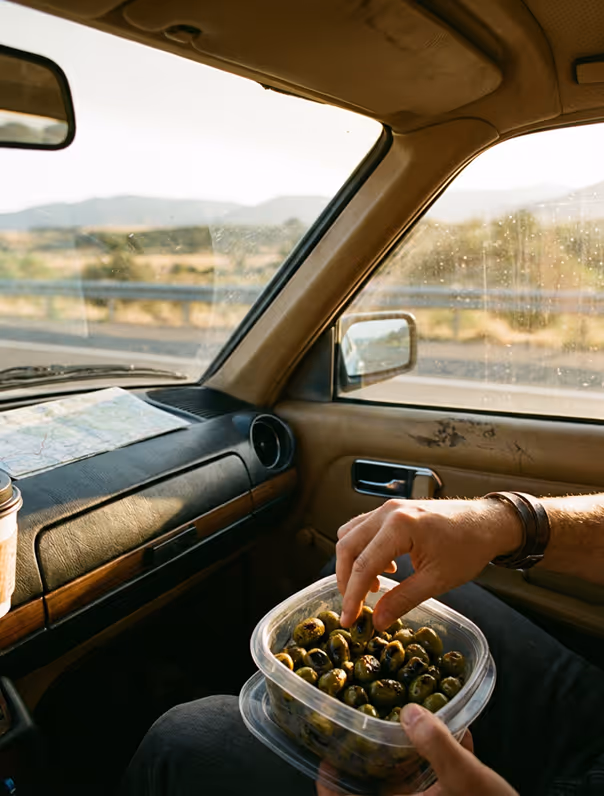Person sitting in car holding a container of olives with one hand picking an olive.
