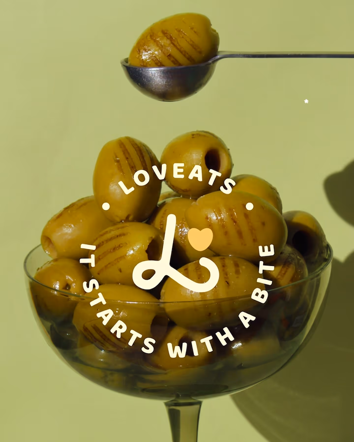 Glass bowl filled with green olives and a spoon holding one grilled olive above it.