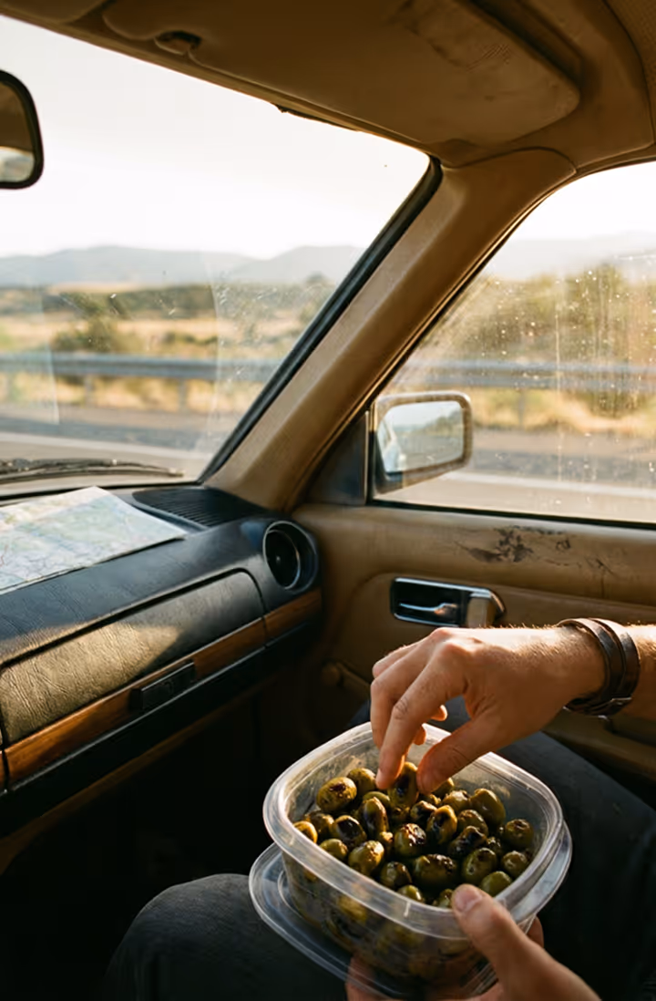 Person picking grilled olives from a plastic container inside a car with a map on the dashboard.