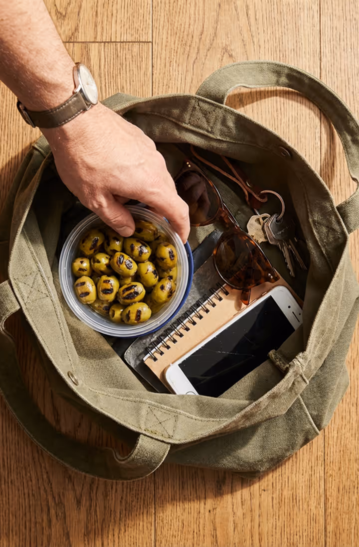 Hand placing grilled green olives in an open green canvas bag with sunglasses, keys, phone, and notebook.