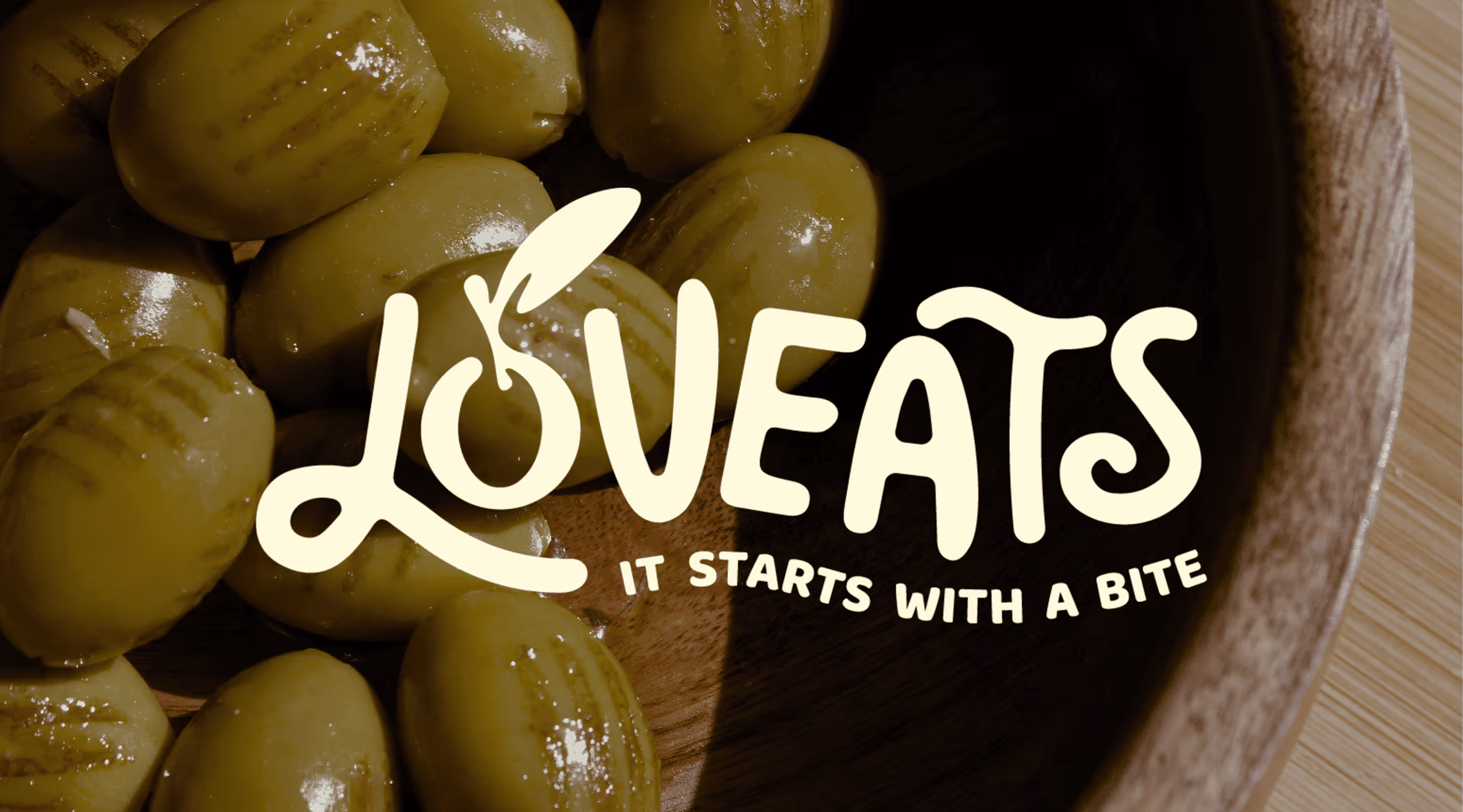 Green olives in a wooden bowl with the text Loveats It Starts With A Bite overlaid.