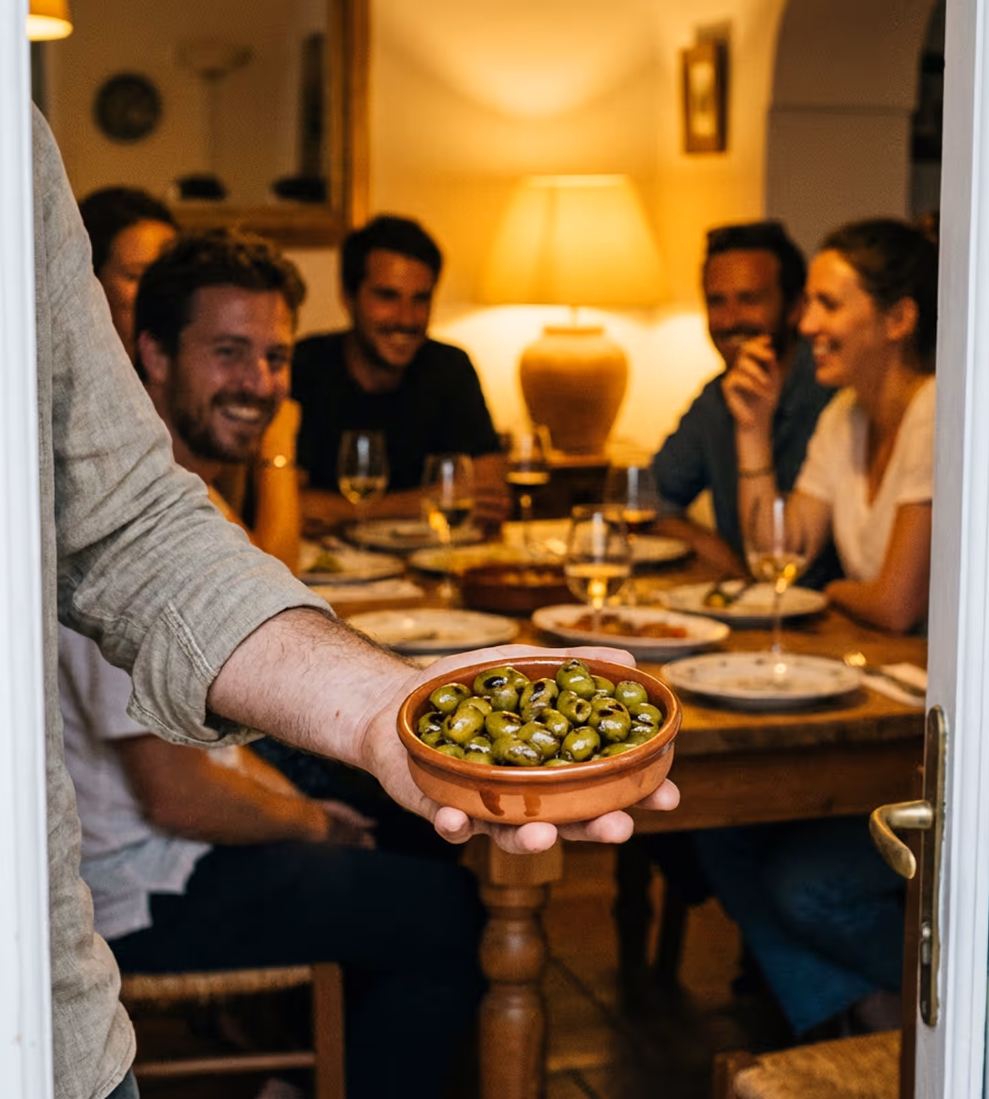 Person holding a bowl of green olives toward a group dining at a warmly lit table.