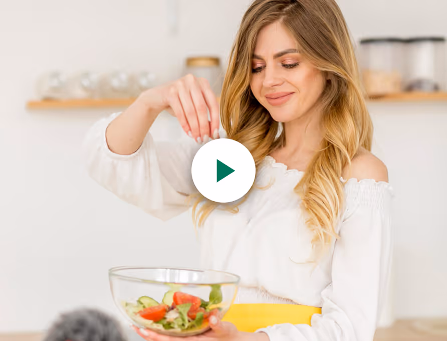 Young woman in a white top holding a glass bowl with salad, sprinkling seasoning over it.