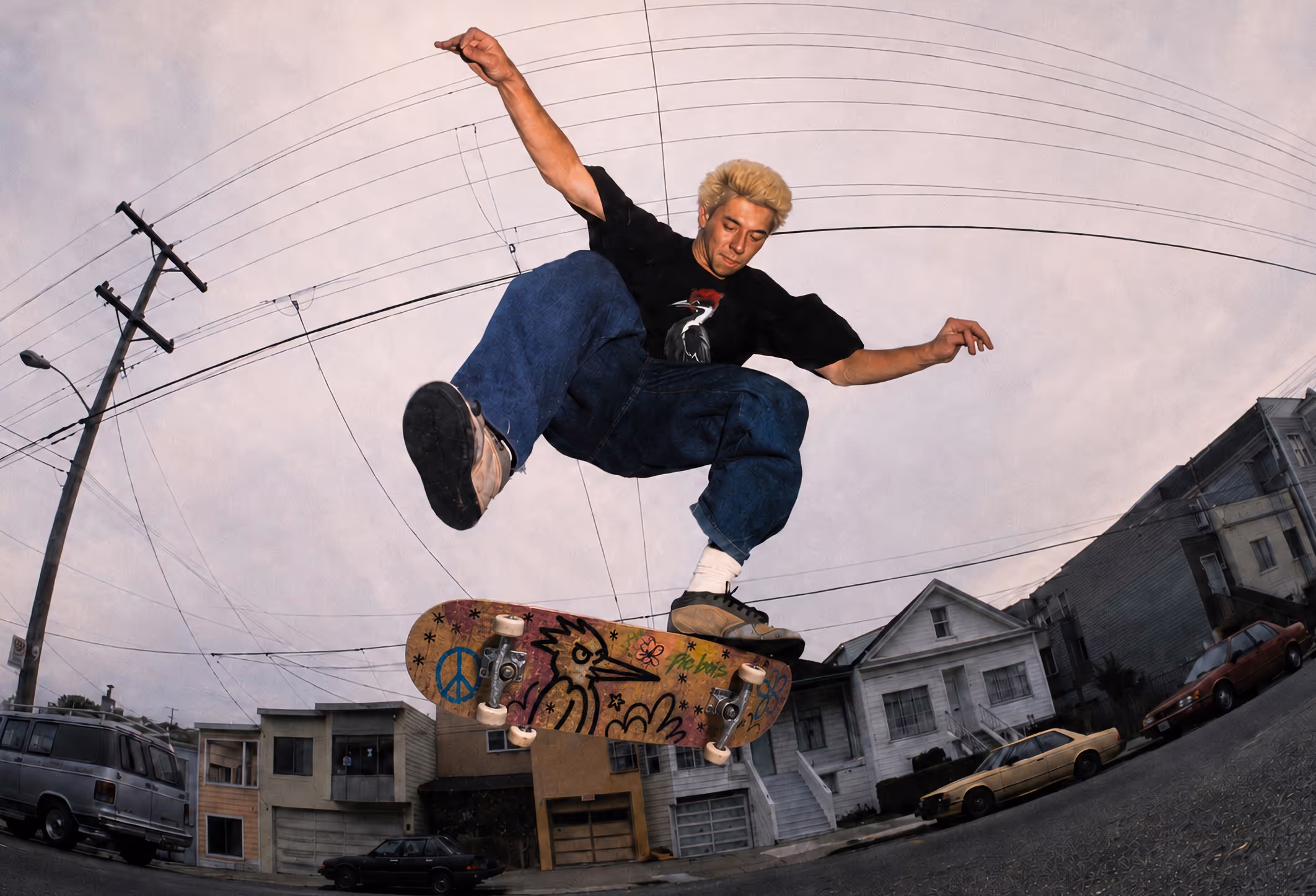 Alt text: Skateboarder performing a trick in an urban neighborhood featuring custom Pic Bois skateboard graphics.  Credit: Composite image using edited photography and custom branding elements.