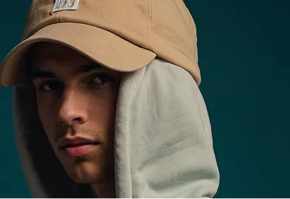 Close-up of a young man wearing a tan cap and a grey hood against a dark background.