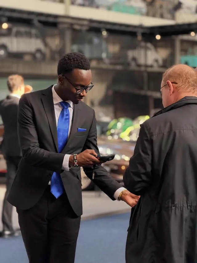 Un homme en costume noir avec cravate bleue vérifie un appareil pour un autre homme en manteau noir.