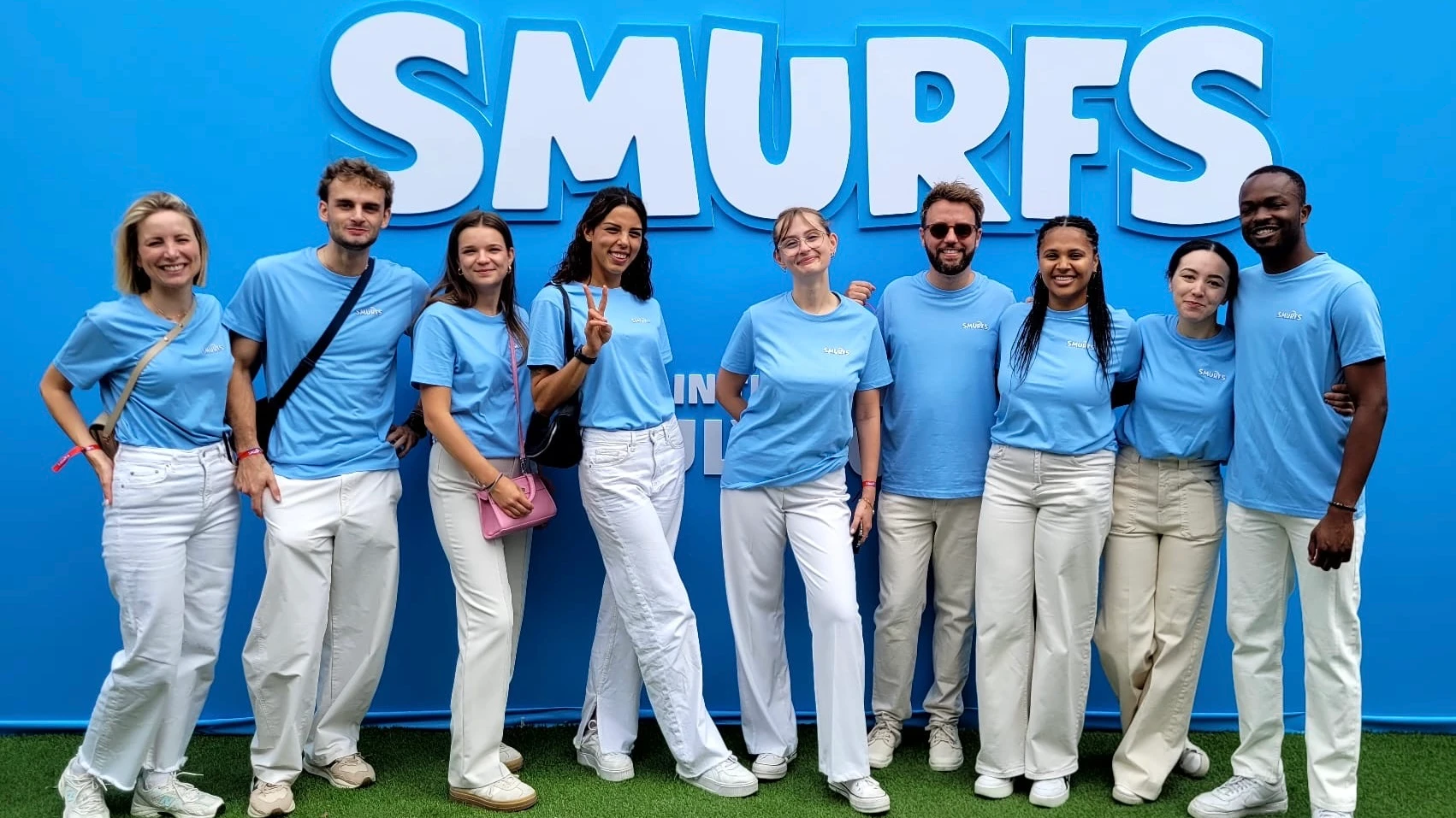 Une équipe de neuf personnes en t-shirts bleus et pantalons blancs pose devant un panneau marqué 'SMURFS'.
