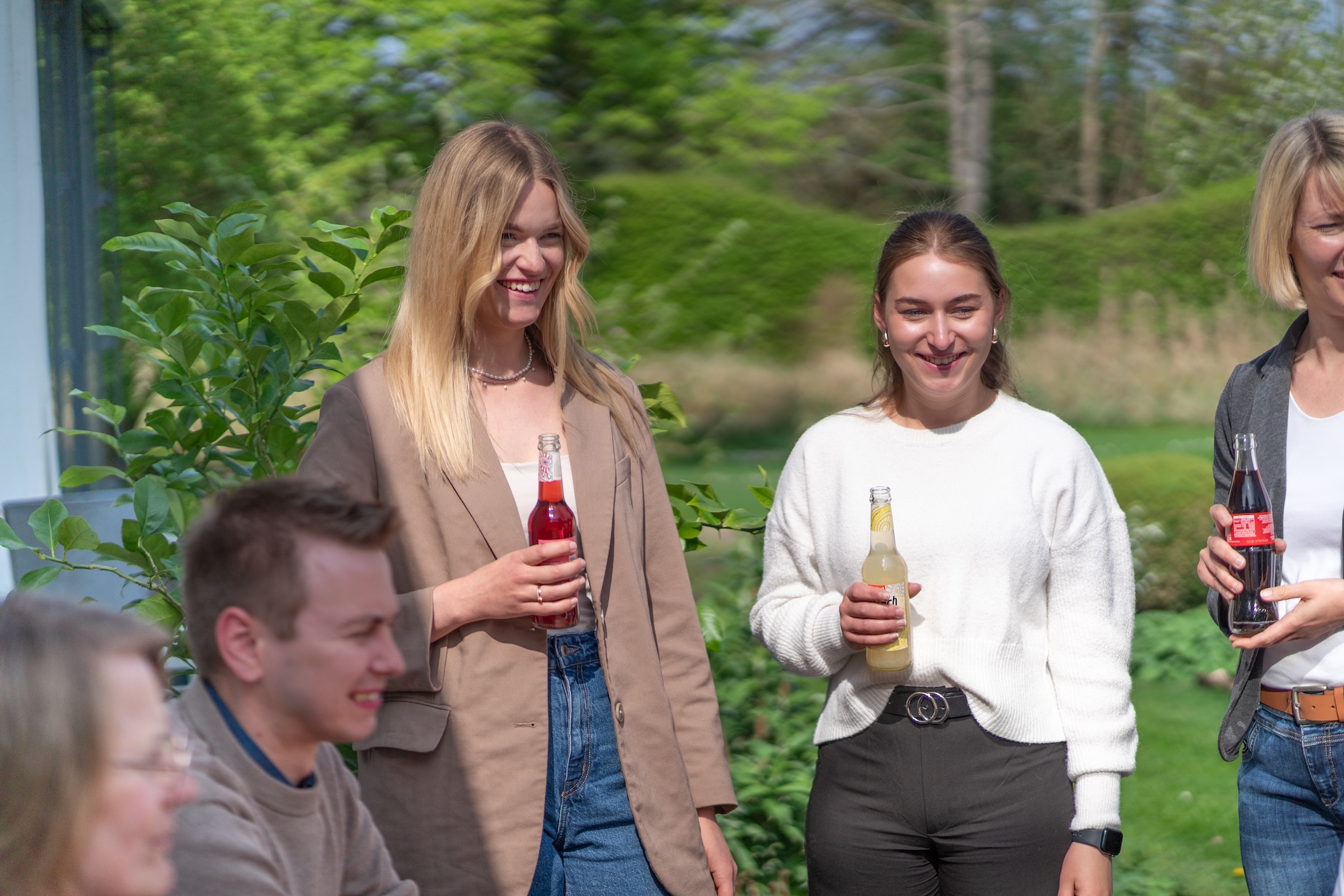 Drei Frauen und ein Mann draußen in einem Garten, die jeweils eine Flasche Erfrischungsgetränk halten und lächeln.